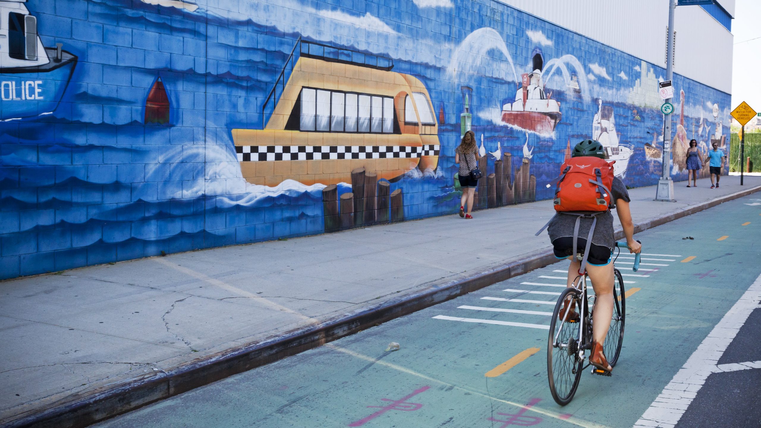 — title missing — Woman biking on bike path next to mural.