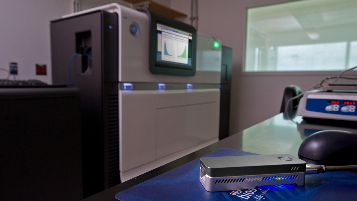 nanopore-sequencer-1200 A small nanopore sequencer (about the size of a short stapler) sits on a table in the foreground. In the background is a large sanger sequencer machine (the size of a large copy machine)