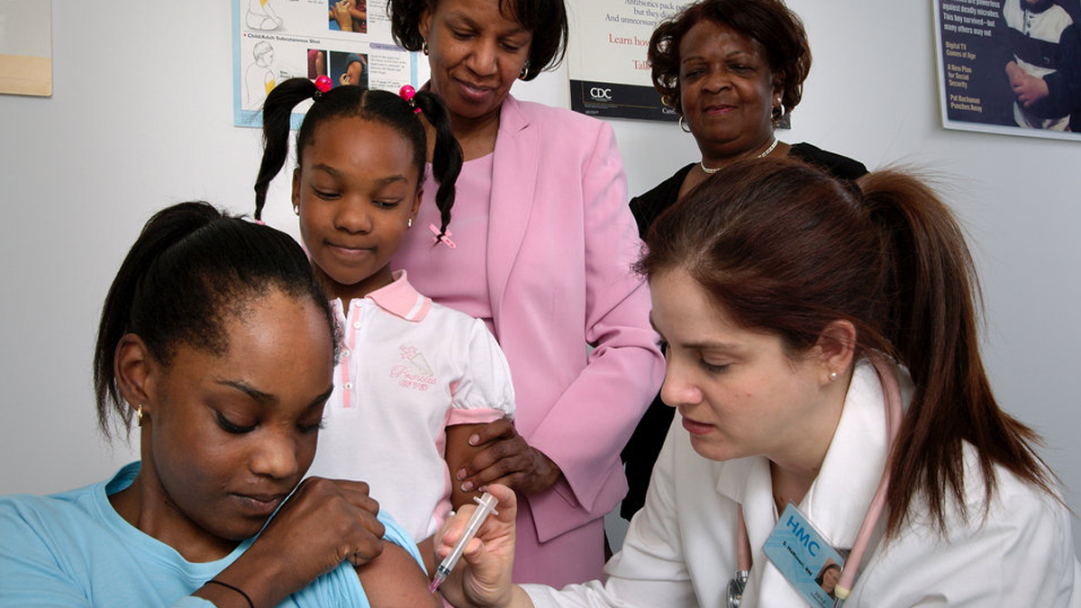 pertussis-1200 A nurse in the process of administering an intramuscular vaccination into a girl’s left arm whose sister, mother, and grandmother watched from the background
