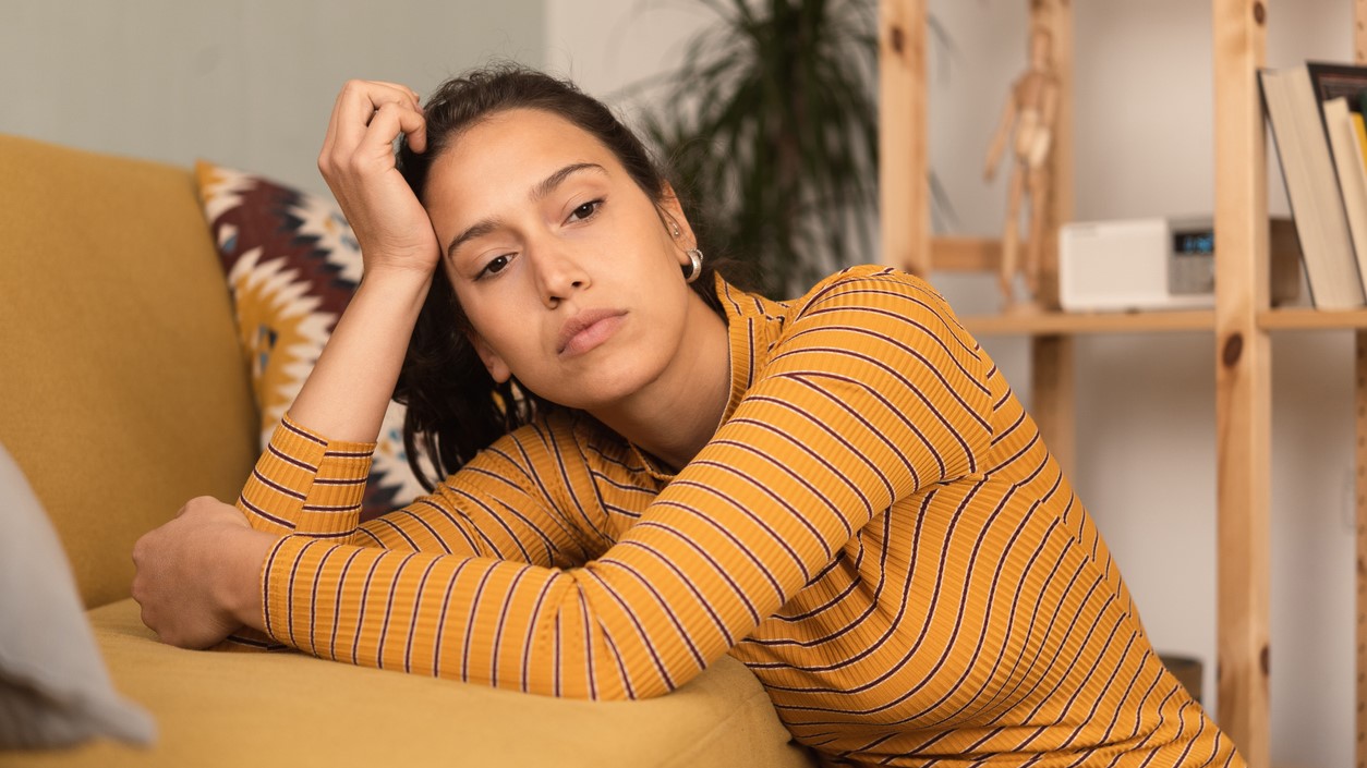 16:9 young woman unwell in home Person not feeling well- sitting on the floor and leaning on couch with their right hand against the side of their head.