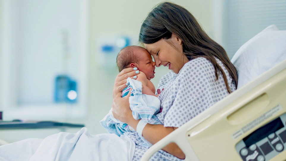 mPINC Survey A new mom sits in a hospital bed and holds her newborn in front of her. She is wearing a hospital gown.