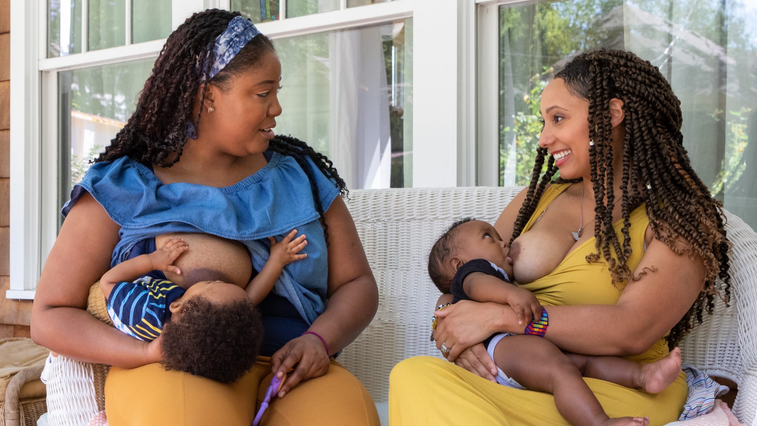 — title missing — Two women breastfeed two children while sitting on a porch outside a house.