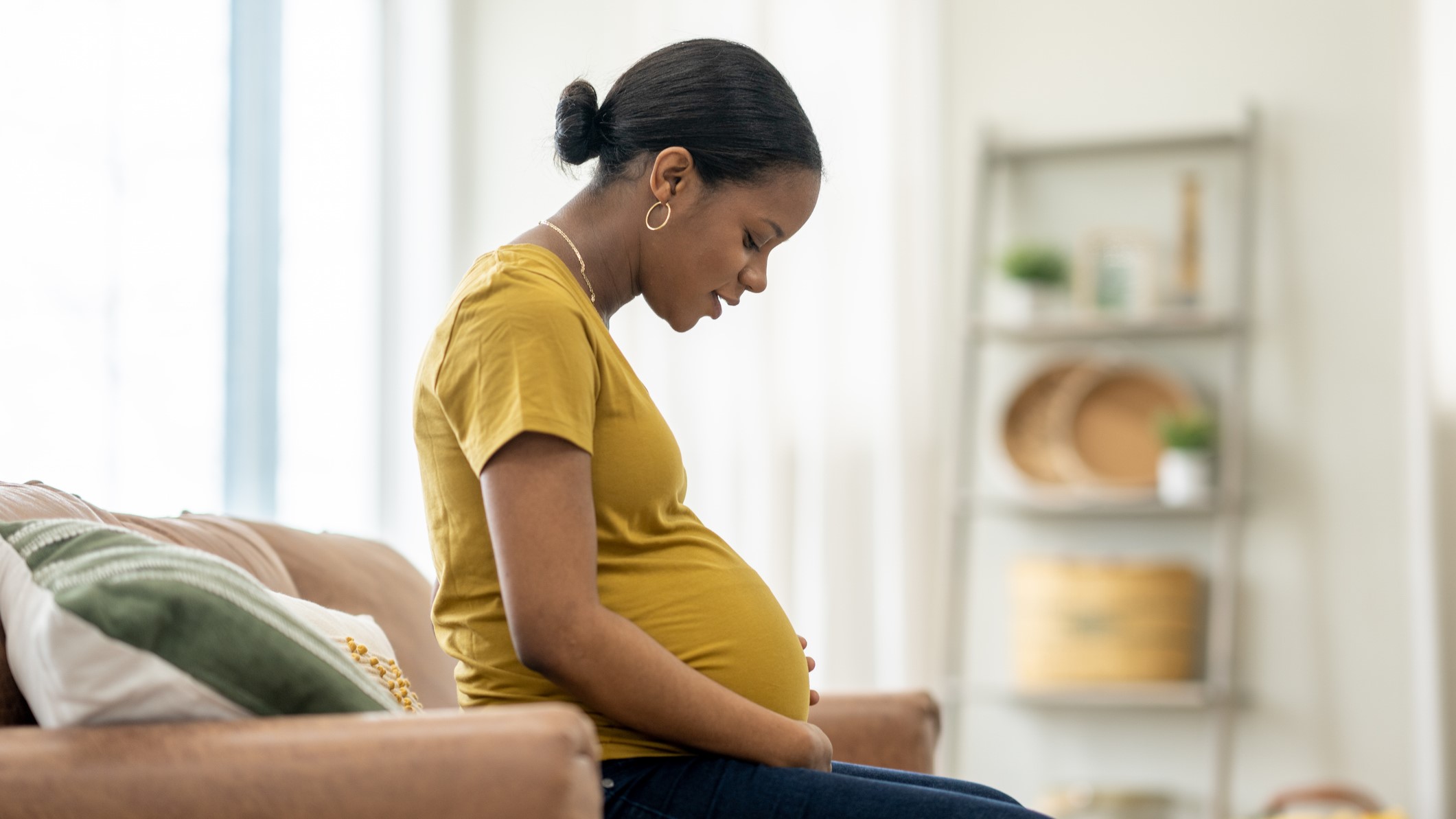 Pregnant Pregnant woman in a yellow shirt sitting on a couch looking at their stomach.