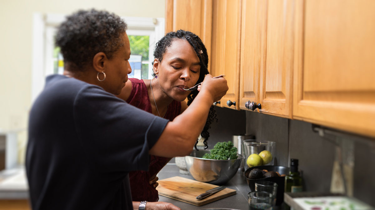 Supporting caregivers Slightly older woman giving a spoonful of what's cooking to younger woman.