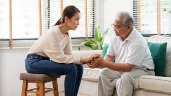 Caregiver sitting and holding the hand of an older person Young woman caregiver sitting on stool holding the hand of an older man on a sofa.