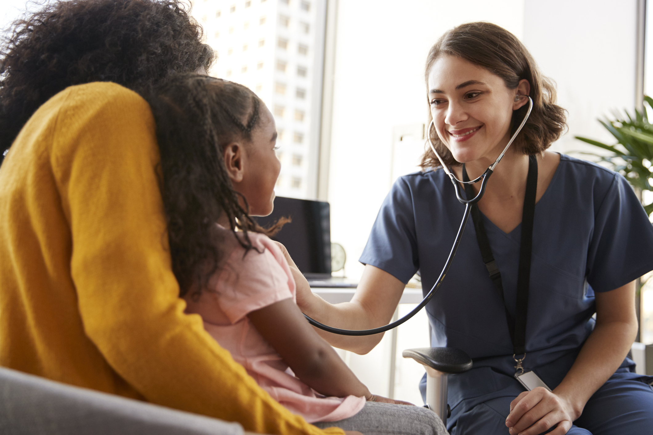 Female Pediatrician Wearing Scrubs Listening To Girls Chest With Stethoscope In Hospital Office Pediatrician using stethoscope on girl on mom's lap.