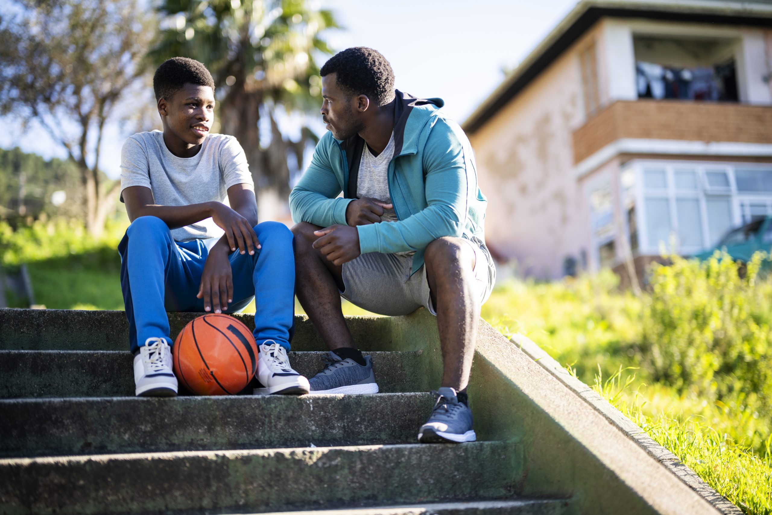 — title missing — A man and a child are sitting and talking on steps outside a housing area. A basketball lies at the feet of the young boy.