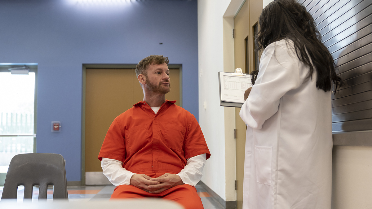 -- title missing -- A man in an orange jumpsuit listens to a person in a white coat with a clipboard.