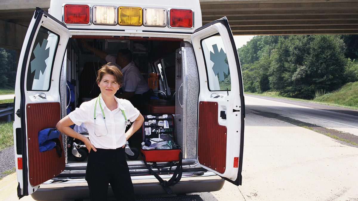 An Emergency Medical Services technician standing outside the back of an ambulance with its doors open.