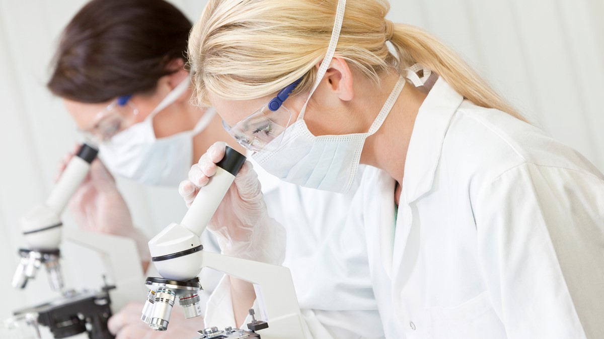 Two lab technicians wearing white lab coats, masks, and protective glasses looking through microscopes.