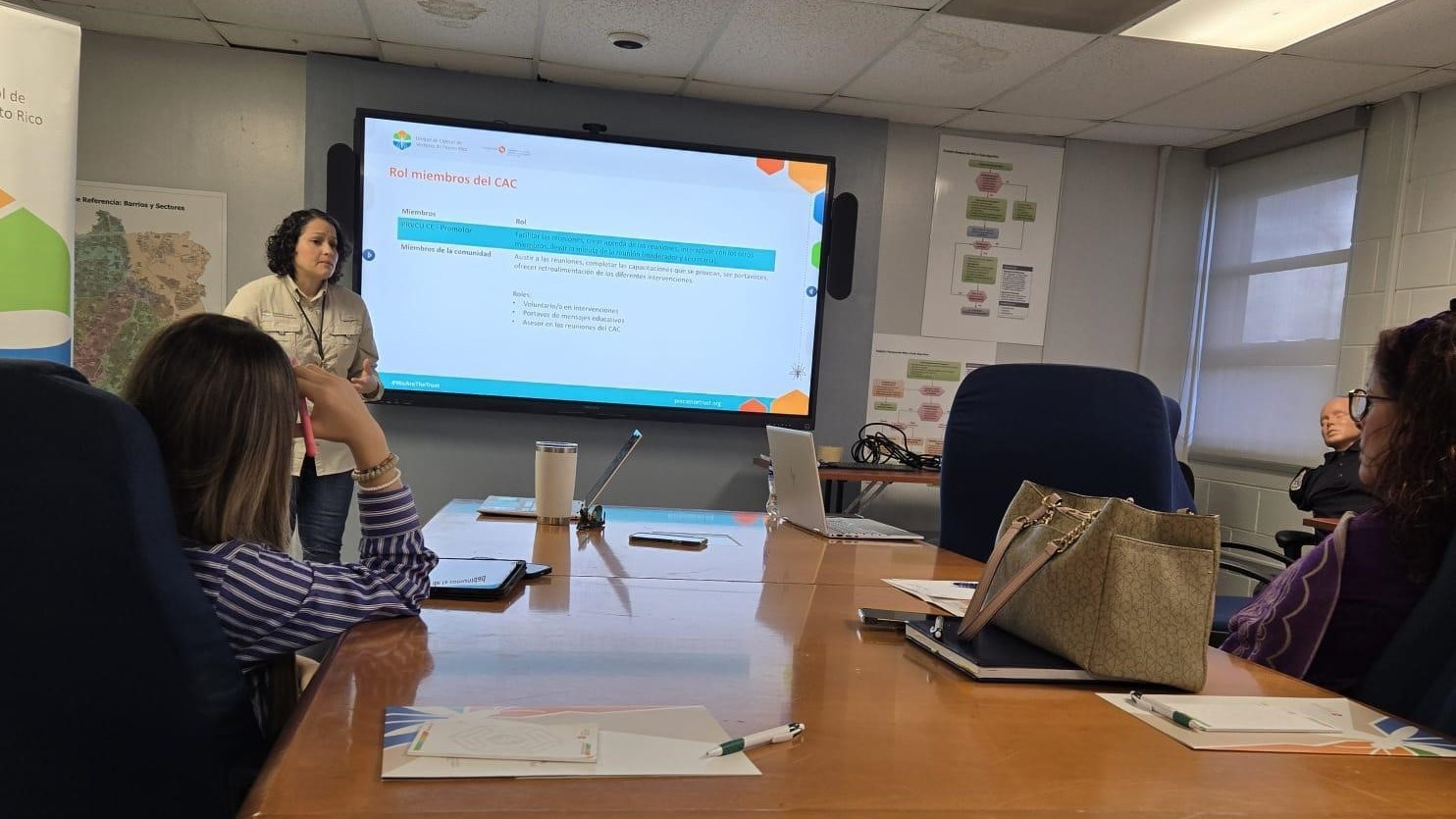 Bayamon IMM CAC A woman presenting in front of a screen while two women sit at a table in the audience.