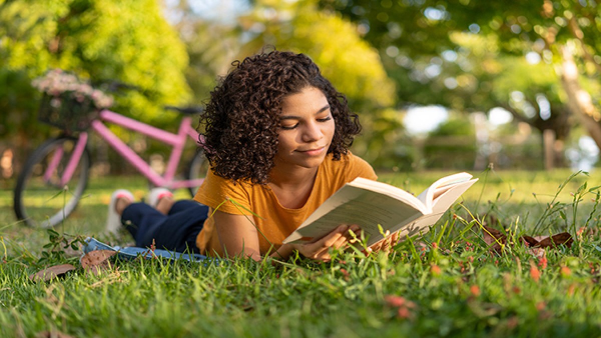 — title missing — Woman reading a book while lying on the ground in a park.