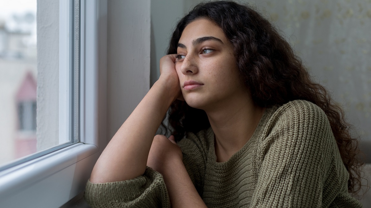 A woman experiencing stress A woman with long, dark hair wearing a green sweater, resting arm on a windowsill, looking out.