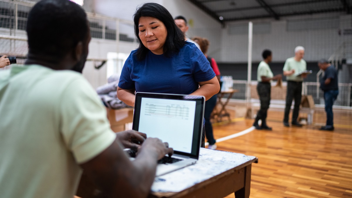 Woman checking in at emergency shelter Volunteer talking with a woman while working on a laptop at an emergency shelter with others in the background.