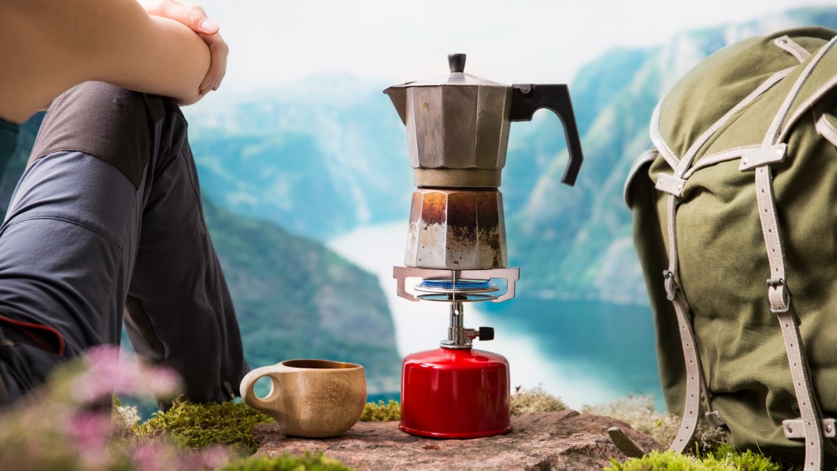 Boiling water hiking in mountains Hiker sitting on a rock looking out over mountains and a water body. A mug, small kettle boiling over a gas burner, and backpack are next to the hiker on the rock.
