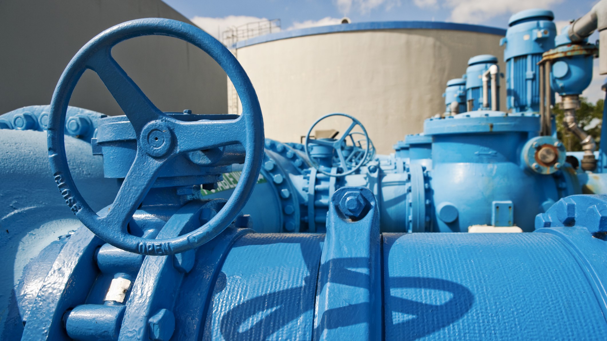 — title missing — Pipes and pumps used to move water at a treatment plant in the foreground, and large water tanks in the background