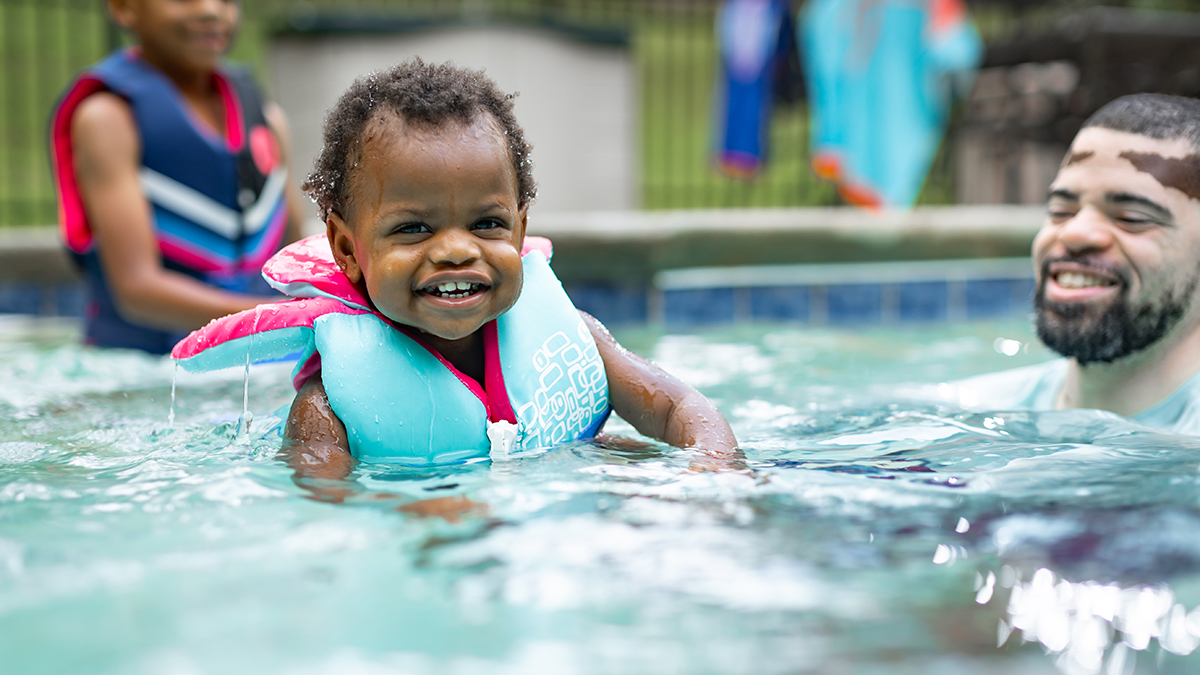 Infant in life jacket w/Dad and family in pool Infant in life jacket with sibling in life jacket in background, together with supervising adult in swimming pool.