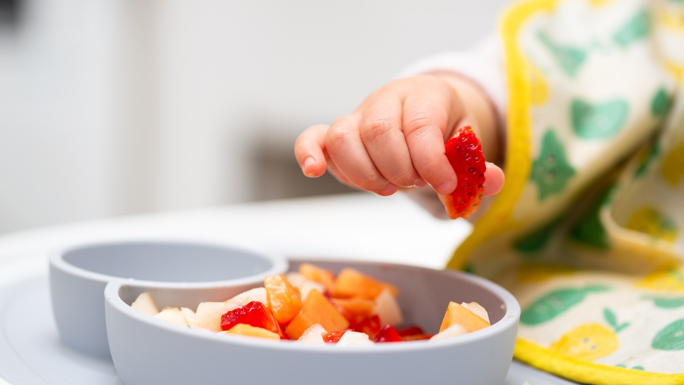 early child nutrition Close up of baby hand with a small piece of fruit over a plate of diced fruit