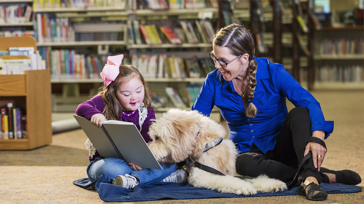 School librarian reading School librarian sitting on the floor reading a book aloud with a young girl and the girl's service dog
