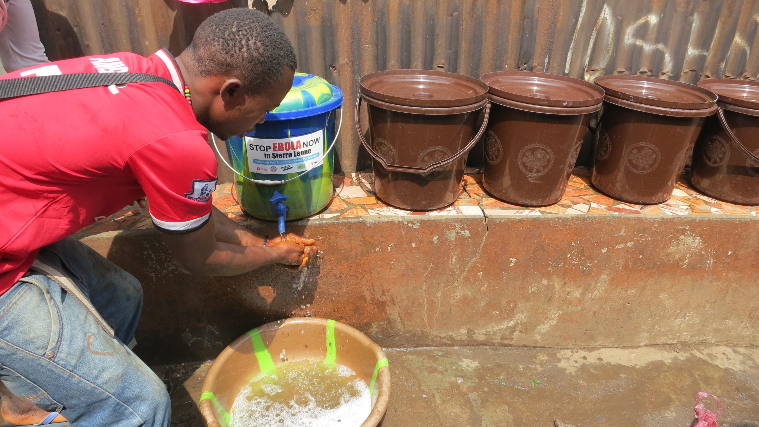 A young man washes his hands using a bucket handwashing station.