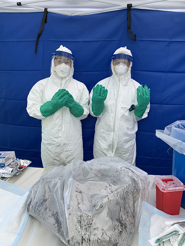 petras-in-ppe Two people in white biohazard suits stand behind a table. On the table is a multi-gallon plastic bag with unknown material inside.