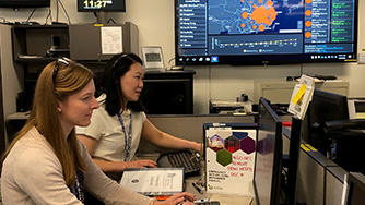 Amy Xie Two women sitting at a desk with a data screen in the background.