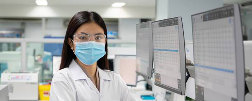 workforcebanner.jpg Masked laboratory worker in lab looking straight ahead with computers that feature data to the side.