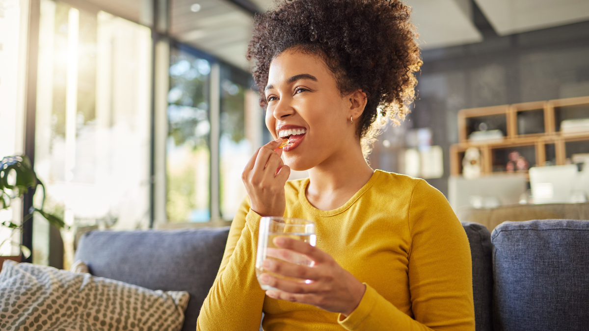 — title missing — Young adult wearing a sweater and taking a vitamin with a glass of water while seating on the couch.