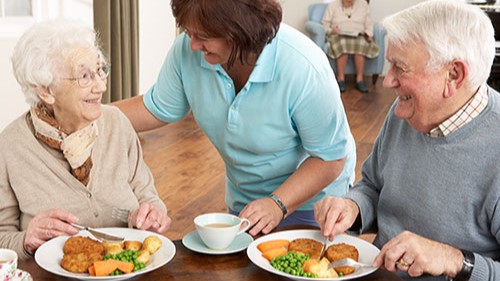 — title missing — Staff member checking on elderly couple in assisted living dining room.