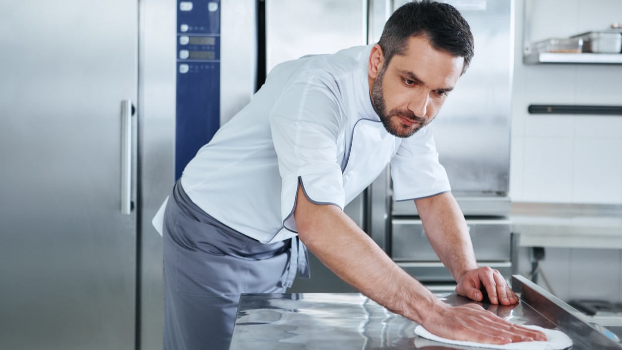 — title missing — Man cleaning worktable in commercial kitchen.