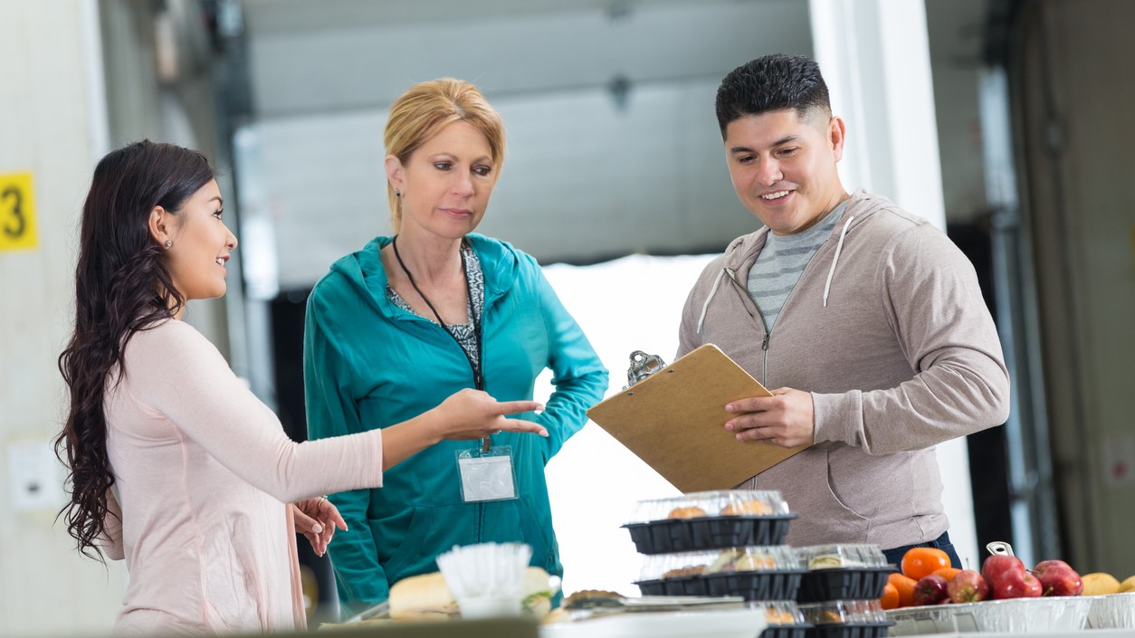 — title missing — Three adults talking by a food service counter. One has a clipboard, and one is wearing a badge.
