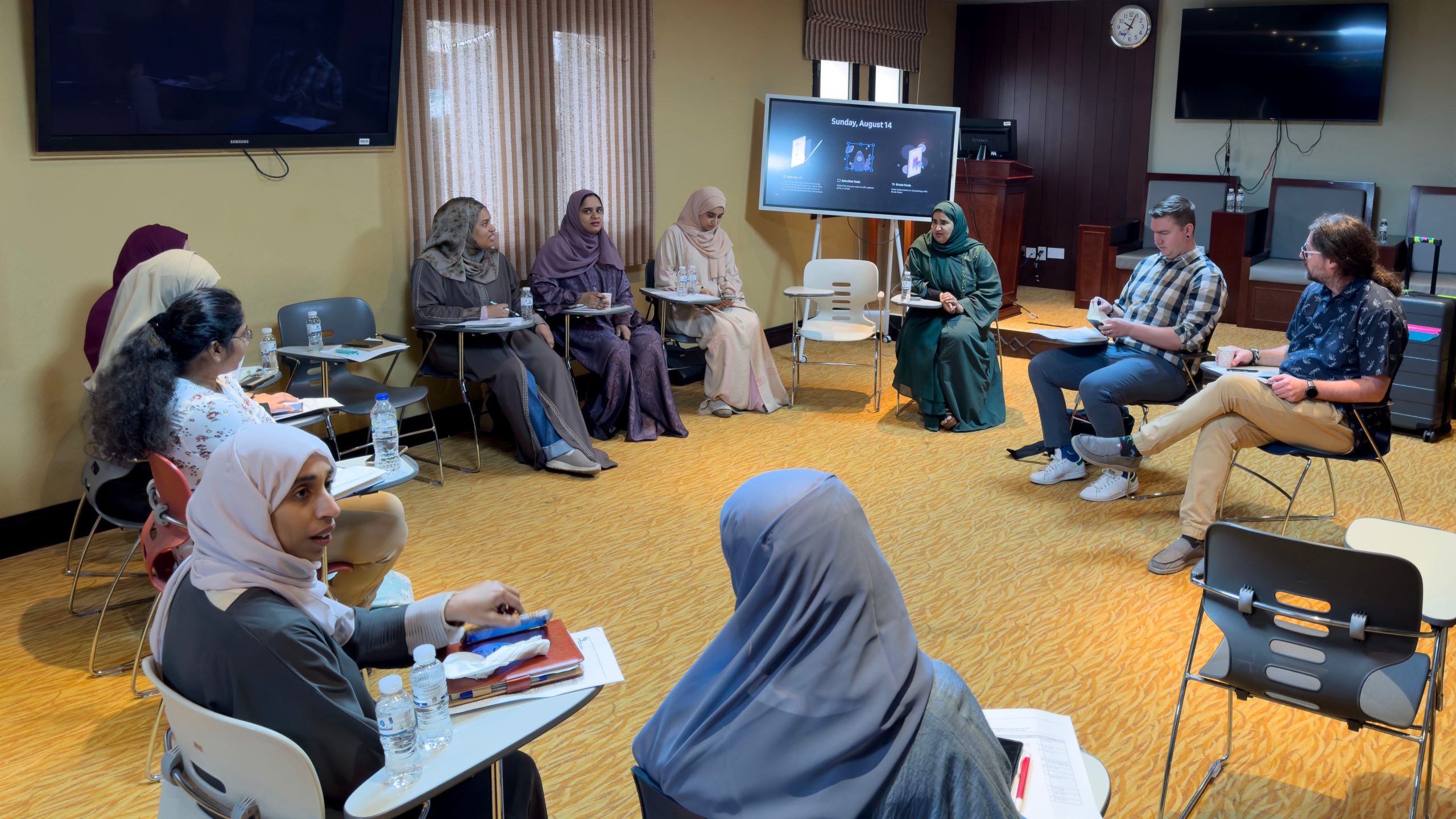 MENA Lab Twinning A group of individuals sitting at desks engaged in conversation with one another.