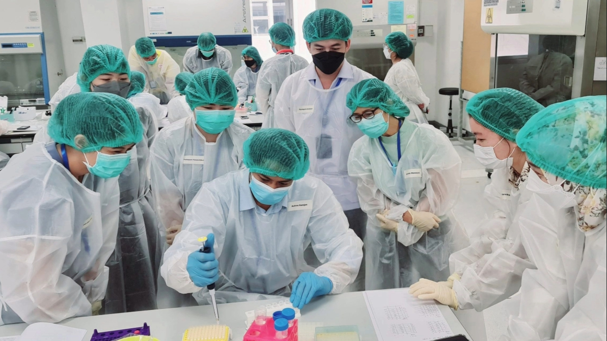 Laboratory_workers.jpg A group of people in personal protective equipment huddle around a table to watch one person who is holding a pipette over a desk.