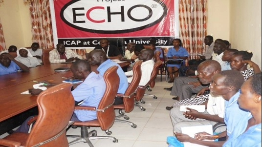SouthSudan_ProjectECHO_Photo A group of people are seated around a table and along a wall. A sign behind them reads "Project ECHO: ICAP at Columbia University."