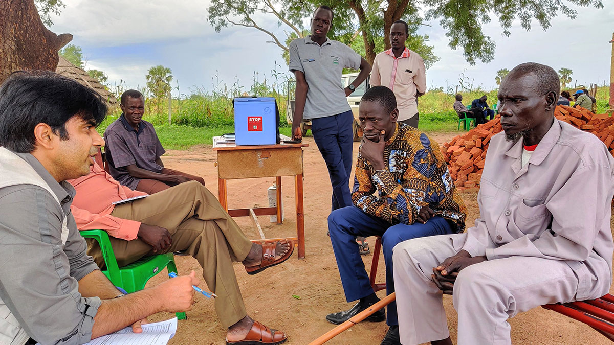 STOP consultant meeting Six men sit across from one another outdoors in front of a tree while two men stand near a table with a blue cooler on top. A man holding a packet of paper and a pen is talking.