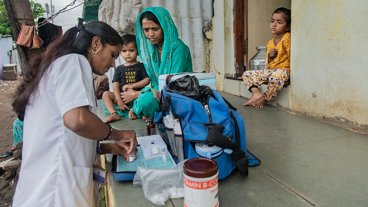 India interventions including polio vaccines and Vitamin A A woman in a white coat twists the lid of a vaccine vial on a porch, where a woman and two children are sitting and where there is a blue bag and a container labeled "vitamin."