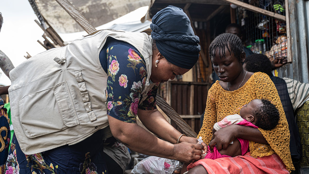 Infant vaccination in Nigeria Outside of a wooden building, a woman wearing a khaki vest and a blue head scarf leans over another woman, who is holding an infant in her lap, and gives the infant a shot in her leg.