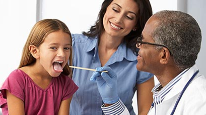 — title missing — A doctor examines a young girl’s throat using a tongue suppressor. Smiling mother in background.