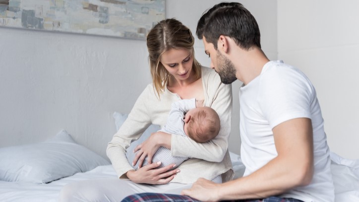 Mother breastfeeding with father support Mother sitting on a bed breastfeeding an infant while the father offers support.