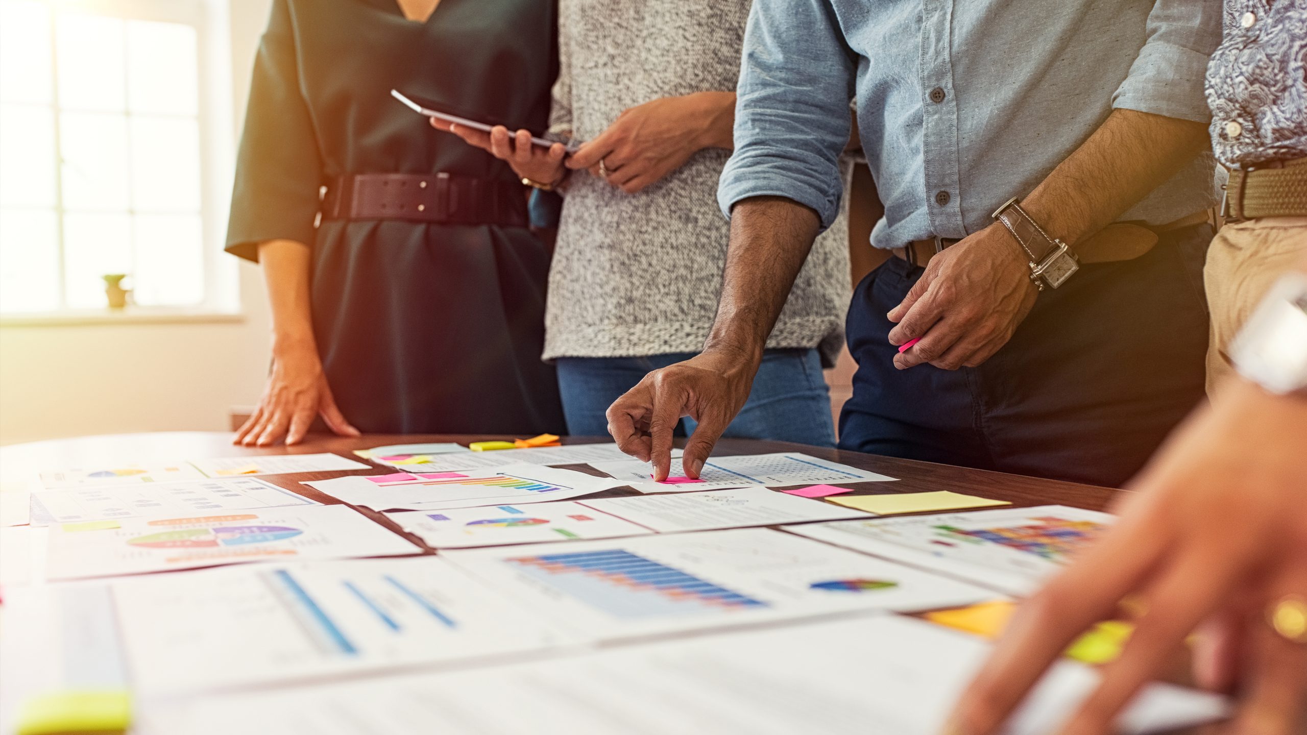 — title missing — A group of people standing around a table with pieces of paper with charts and graphs.
