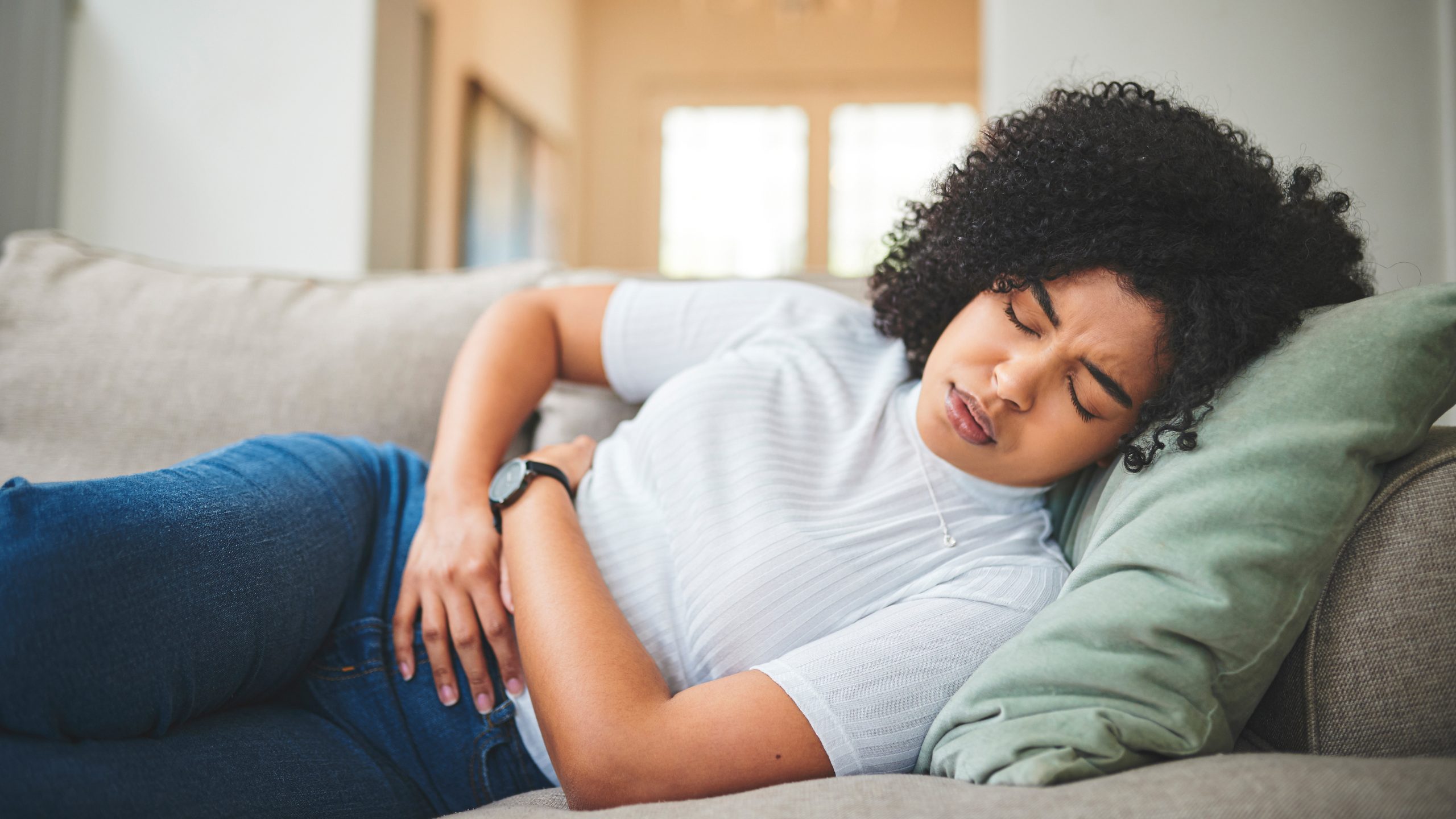 A young woman lays on a couch holding her stomach in pain
