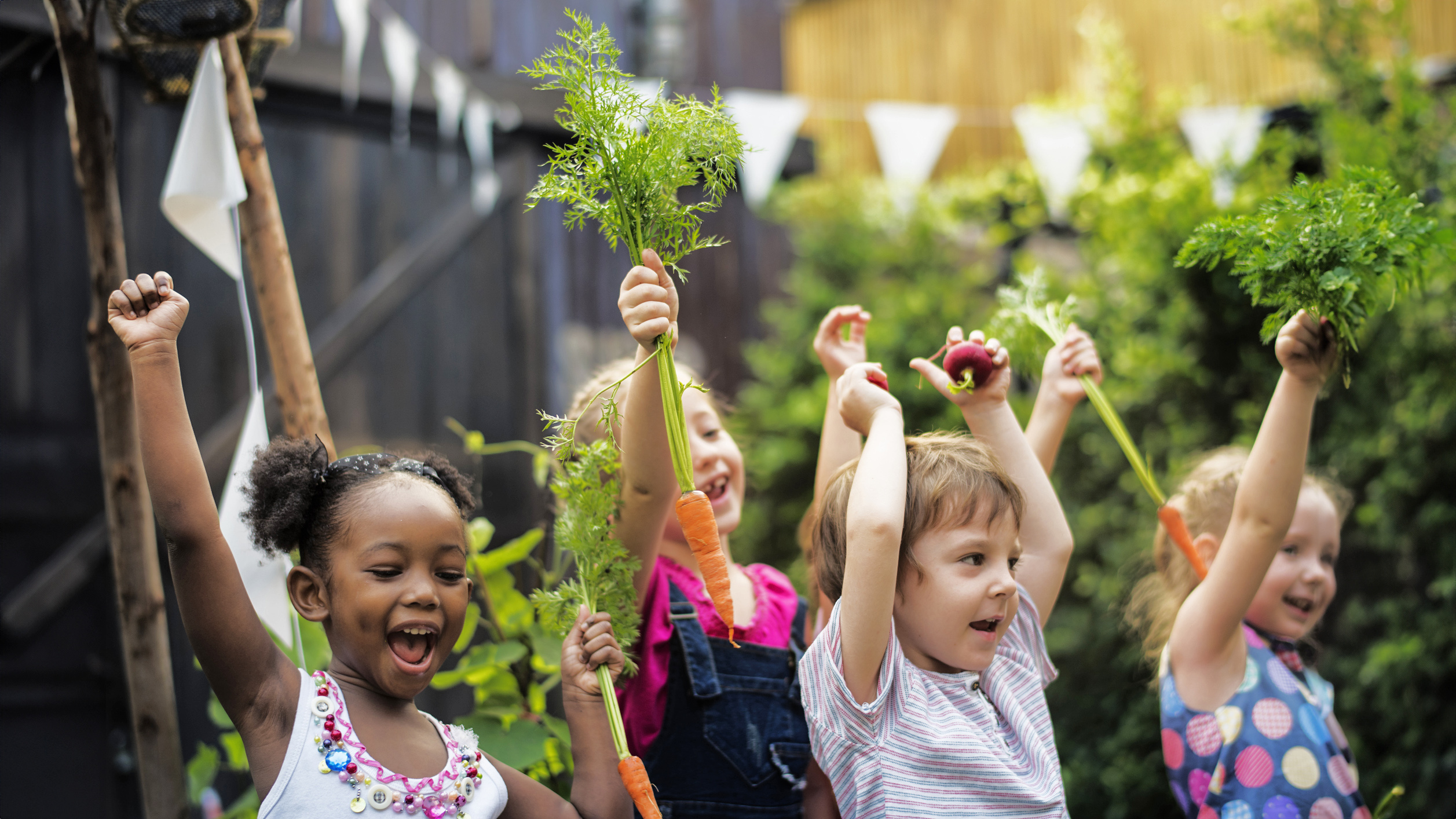 Kids in a vegetable garden with carrot Children in a garden, with their hands up in the air, holding picked greens.