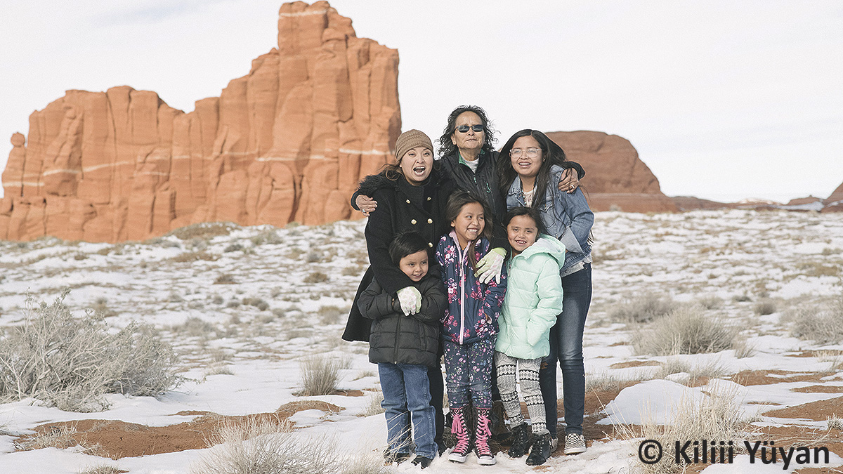 Healthy Tribes Navajo Family and Rocks - credited Older American Indian woman hugging two younger women and three younger children in front of them.
