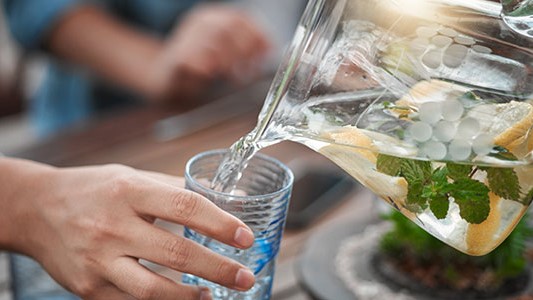 — title missing — A hand pouring water from a pitcher with fruit and mint into a cup.