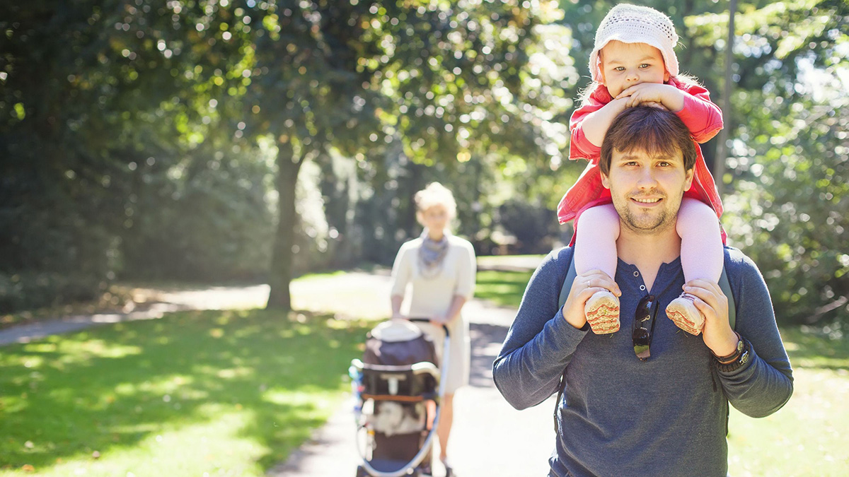 Family of four Man walking in the park with toddler on his shoulders, and a woman behind him walking with a stroller