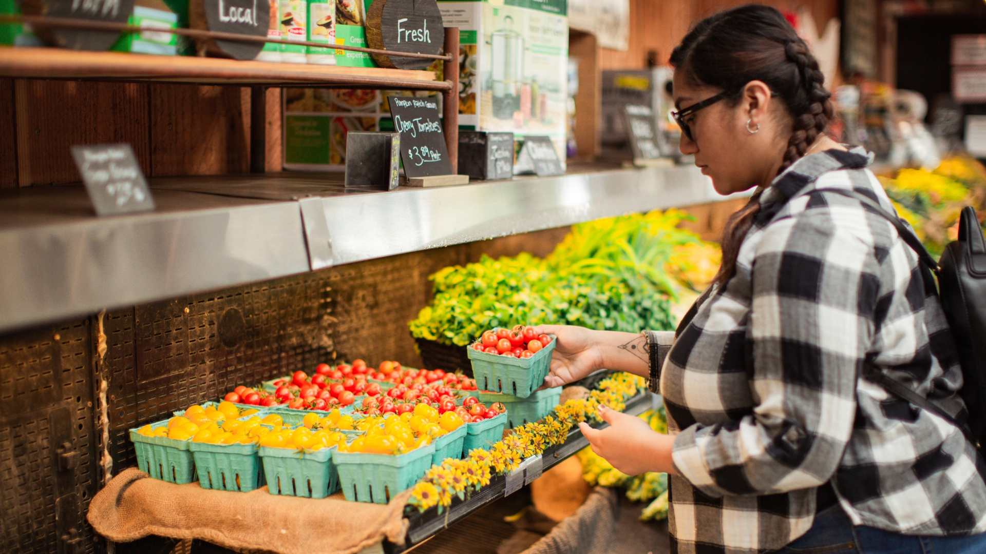 Woman Grocery Shopping Pregnant Hispanic woman wearing a black and white checkerboard shirt, jeans, glasses with her hair braided. She is holding a carton of cherry tomatoes from the produce section at a local farm store.