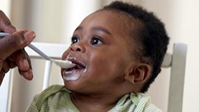 Introducing solid foods 6 month old baby sitting in high chair and being fed solid foods for the first time from a spoon.