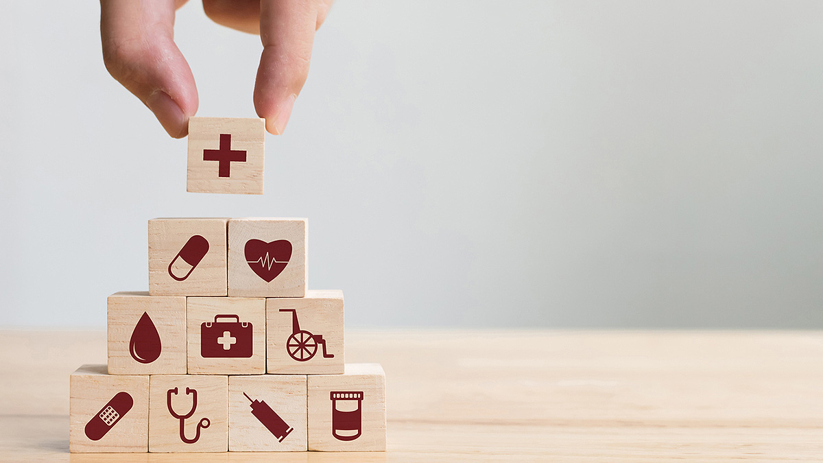 IBD Stacked Medical Blocks Hand placing a final wooden block on top of a pyramid of blocks, each with a different medical symbol on it.
