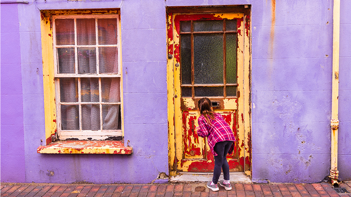 Young girl in front of wooden door A young girl is peeping through a window in a door with peeling and chipping paint.