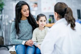 alt="Mother And Daughter At The Doctor's office." A woman and a child sit on a doctor's examine table in front of a doctor.